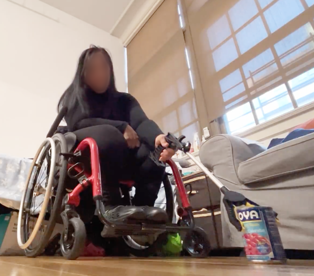 Photograph of a woman wearing all black and sitting in a red wheelchair, using a reacher-grabber tool with an outstretched arm and grabbing a can of Goya kidney beans.