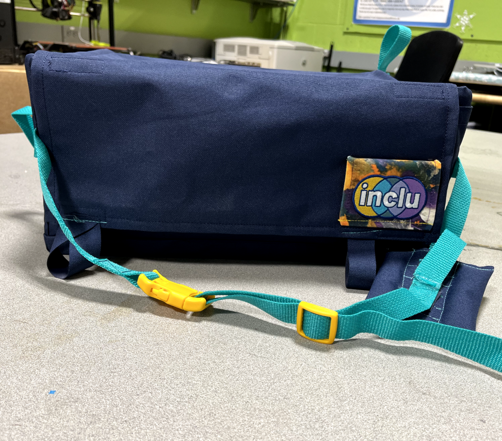 Photograph of the LapSnap assistive device. A rectangular shaped vessel made out of navy blue heavy duty cloth, with teal blue straps and yellow buckles. There is a patch with the company's logo in the bottom right corner of the LapSnap, which reads inclu on a patterned fabric.