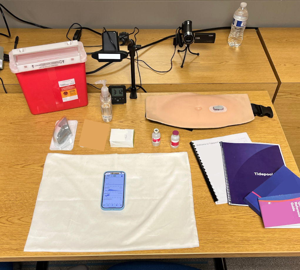 Human factors testing setup of several items on a desk underneath a webcam. The items include a sharps container, an iphone, several paper booklets, insulin vials, hand sanitizer, and a CGM sensor and transmitter.