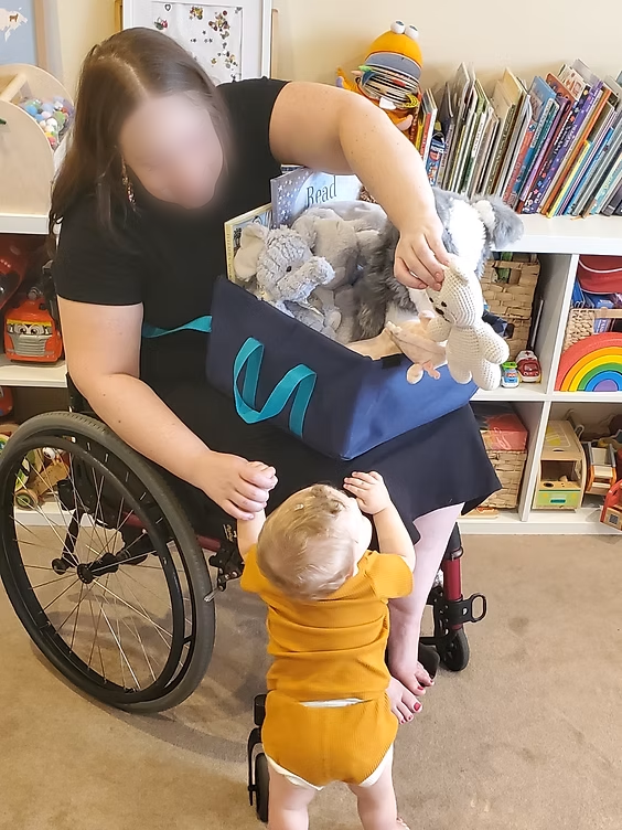 Photo of a woman, wiwth her face blurred for privacy, sitting in a wheelchair with a blue lapsnap basket full of children's toys in her lap and reaching over the side of her chair to hold a baby's hand.
